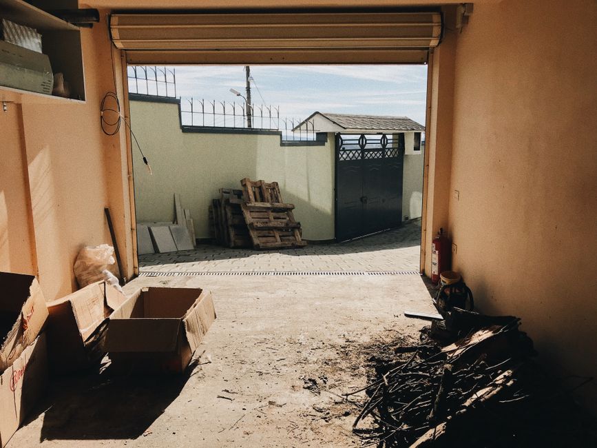 A person with curly black hair, wearing a black T-shirt and plaid shorts, is standing next to an open-top skip filled with a substantial load of broken wooden planks, panels, and debris. The wooden ma