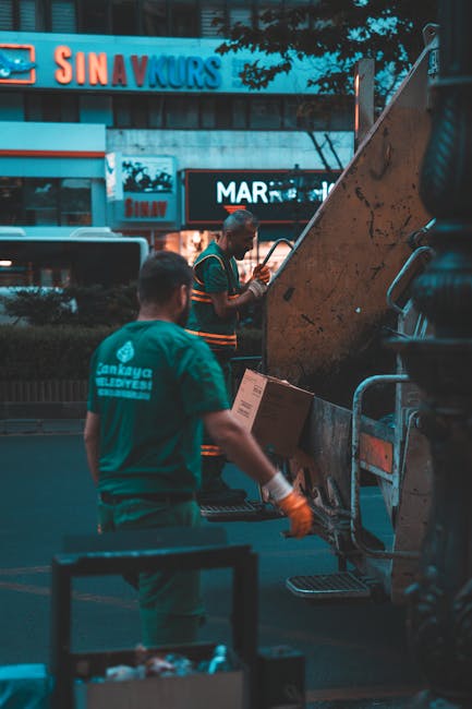 Two workers in teal uniforms with rubber gloves are operating a waste collection vehicle in an urban setting during the evening. One worker, standing closer to the camera, faces away and holds a clipb