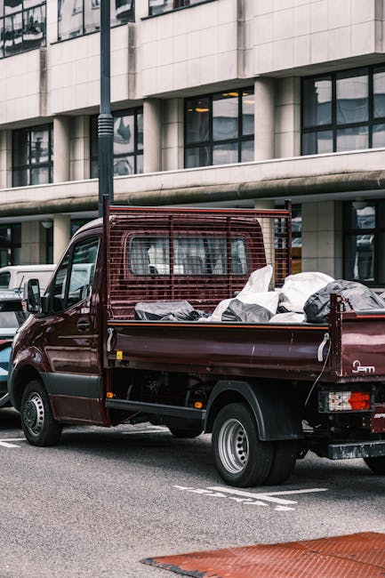 A waste collection worker dressed in a green uniform with reflective orange stripes is standing next to the open rear compartment of a small commercial rubbish truck on a tree-lined urban street. The 