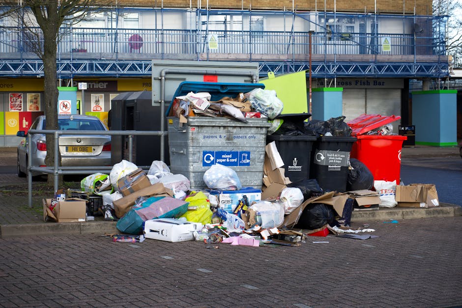 A collection of overflowing waste and rubbish bins is situated on a paved sidewalk near a parking area, with various types of discarded materials spilling onto the ground. The scene includes large gre