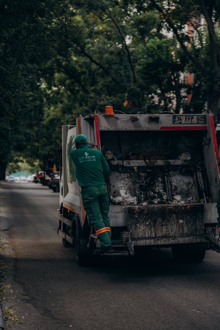 A collection of overflowing waste and rubbish bins is situated on a paved sidewalk near a parking area, with various types of discarded materials spilling onto the ground. The scene includes large gre